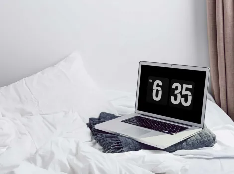 Laptop mit großer Zeitanzeige auf Bett mit weißer Bettwäsche